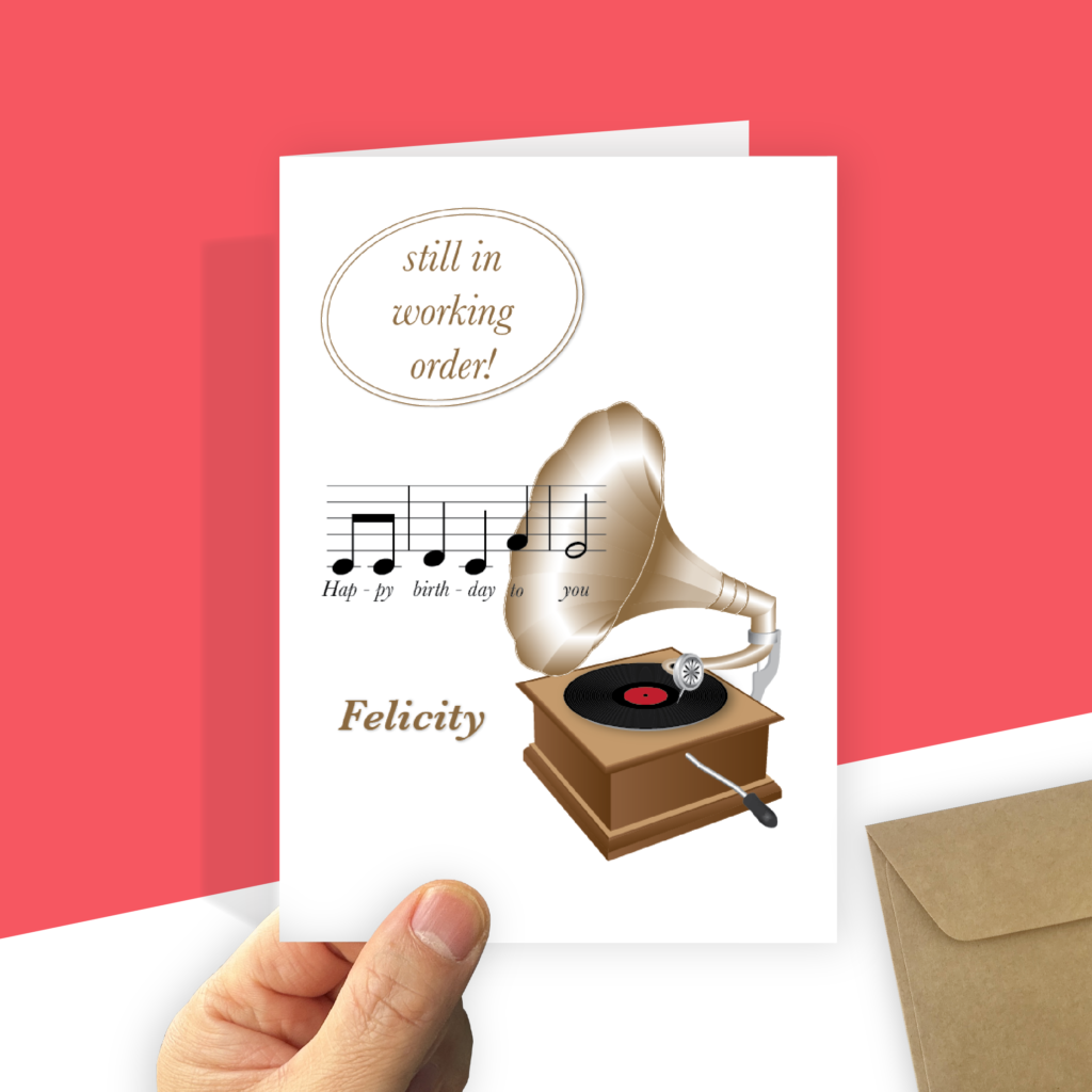 A portrait-oriented birthday card featuring a vintage gramophone with a golden horn and a black vinyl record. Coming out of the speaker, a musical staff displays the notes and lyrics for "Happy birthday to you." At the top of the card, a golden oval frame contains the text "still in working order!" in a classic serif font. Your recipient's name is printed in golden italic font at the bottom left. The card is held by a hand with a recycled brown envelope visible in the corner.