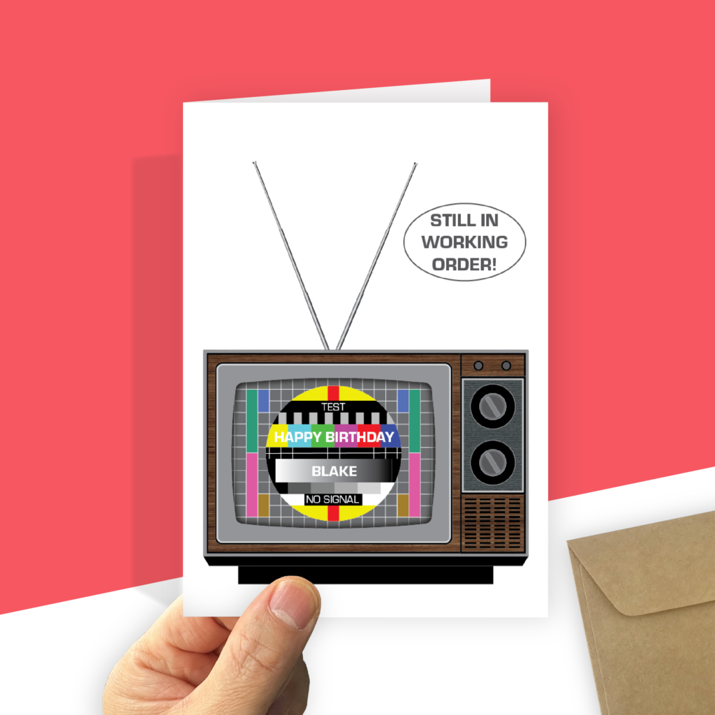 The birthday card features a 1960s TV on a white background. The screen displays a common PAL colour test pattern with white "HAPPY BIRTHDAY" across the colour band. On the gradient black band is your recipient's name also in white. Two silvery aerials are extended in a vee. Above the TV is a bubble that reads "STILL IN WORKING ORDER." A hand holds the card next to a recycled brown envelope.